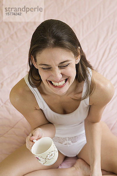 Blick von oben auf eine junge Frau  die auf einem Bett sitzt und eine Teetasse hält.