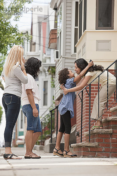 Kind begrüßt eine Frau mit einer Umarmung vor einem Haus  während zwei Frauen zusehen