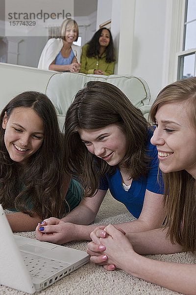 Drei Mädchen im Teenageralter benutzen einen Laptop  während ihre Mütter ihnen über die Schultern schauen