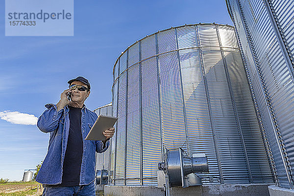 Ein Landwirt steht neben Getreidelagern und benutzt ein Mobiltelefon und hält ein Tablet in der Hand; Alberta  Kanada