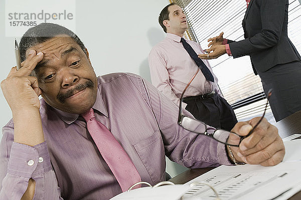 Geschäftsmann  der in einem Büro sitzt und gestresst aussieht  während sich seine Kollegen im Hintergrund unterhalten