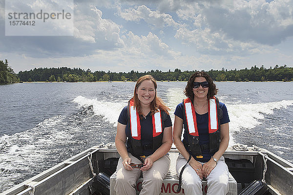 Zwei Frauen in Schwimmwesten sitzen in einem Motorboot und schauen in die Kamera  während es über einen See fährt