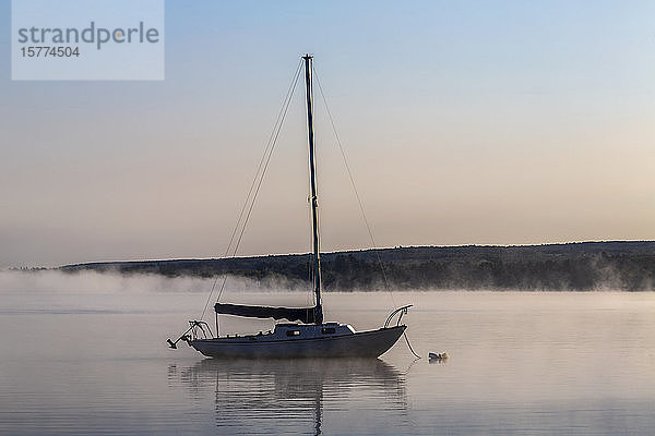 Ein Segelboot liegt auf dem ruhigen  nebelverhangenen See bei Sonnenaufgang vor Anker; Brome Lake  Quebec  Kanada
