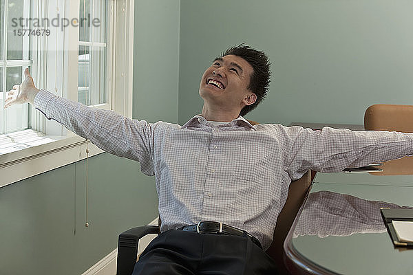 Junger asiatischer Geschäftsmann sitzt auf einem Stuhl am Arbeitsplatz und feiert einen Sieg
