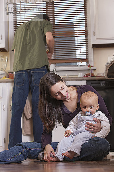 Eine junge Mutter hält ihren kleinen Sohn  während der Vater in der Küche im Hintergrund steht.