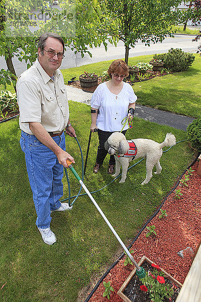 Eine Frau mit einer Behinderung zu Hause bei ihrem Mann  der mit Gehhilfen und einem Diensthund im Hof steht