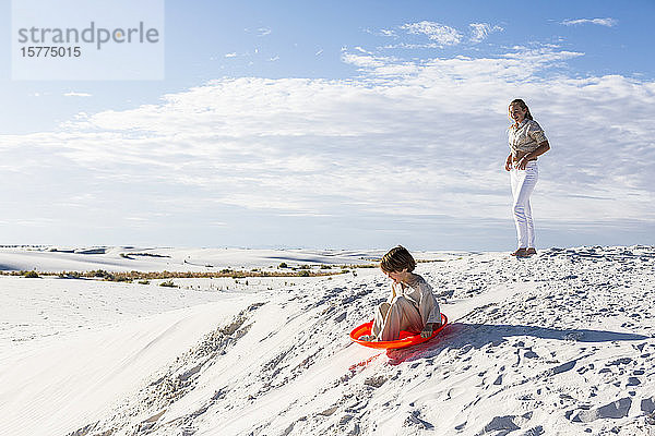 Kinder spielen in einer Sanddünenlandschaft  eines auf einem orangefarbenen Schlitten.