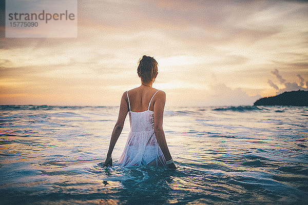 Rückansicht einer jungen Frau in einem weißen Kleid  die bei Sonnenuntergang im Meer spazieren geht.