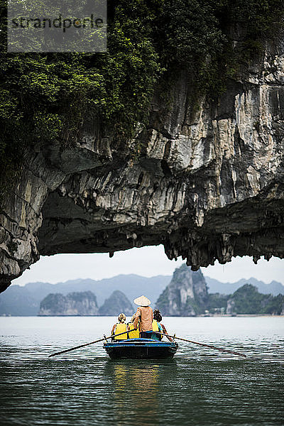 Rückansicht eines Mannes mit Strohhut  der eine kleine Gruppe von Menschen auf einem Boot transportiert und unter einem natürlichen Felsbogen rudert.