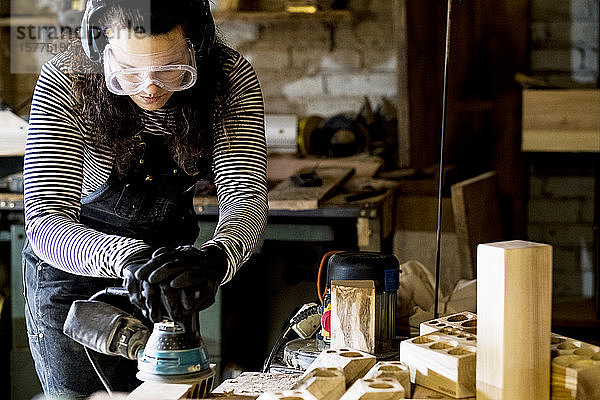 Frau mit langen braunen Haaren mit Latzhose  Schutzbrille und Gehörschutz  die in einer Holzwerkstatt steht und eine Schleifmaschine benutzt.