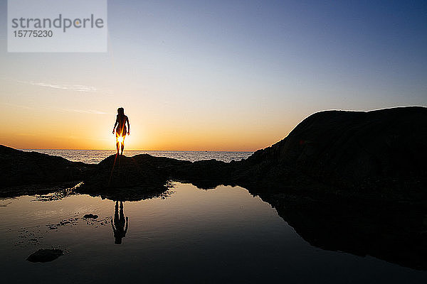 Silhouette einer Frau  die bei Sonnenuntergang auf einem Felsen am Meer steht.