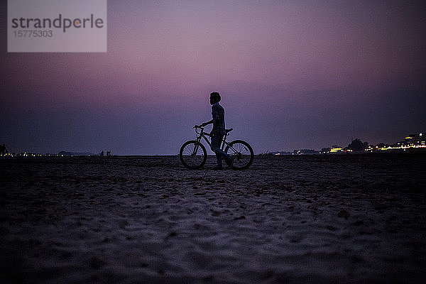 Silhouette eines Jungen  der bei Sonnenuntergang am Ufer eines Flusses ein Fahrrad schiebt.