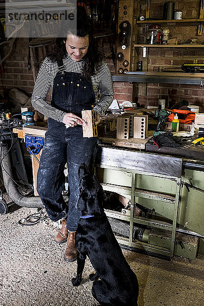 Frau mit langen braunen Haaren in Latzhose in Holzwerkstatt mit schwarzem Labrador.