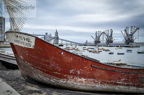 Ein altes Boot mit einem roten schäbigen Rumpf strandete am Ufer am Wasser in Lissabon. In der Ferne Schiffskräne und eine große moderne Straßenbrücke.