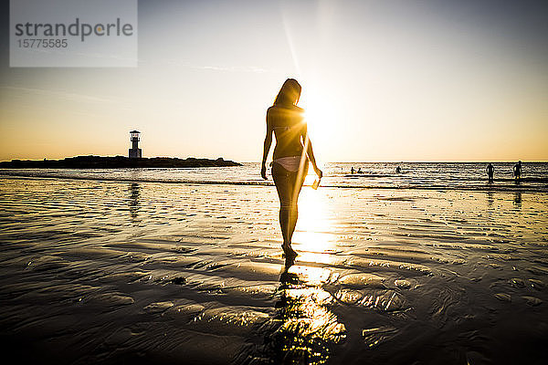 Rückansicht einer Frau  die bei Sonnenuntergang in Richtung Strand geht.