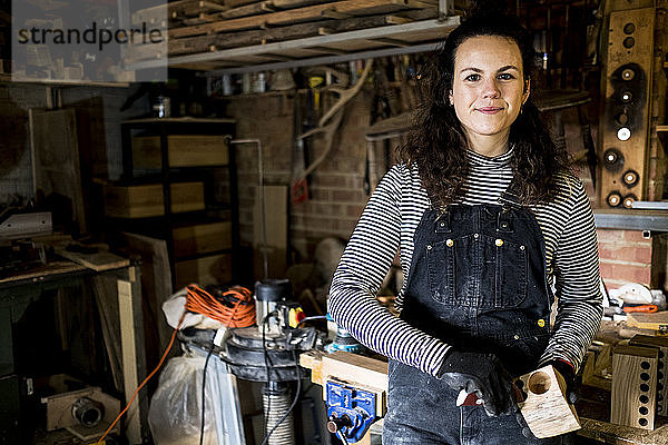 Frau mit langen braunen Haaren in Latzhose  steht in Holzwerkstatt und lächelt in die Kamera.