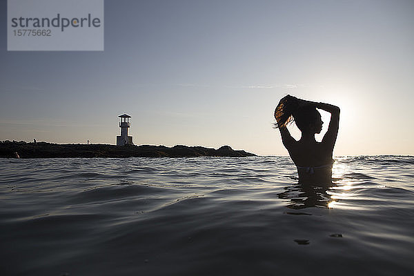 Rückansicht einer im Meer badenden Frau bei Sonnenuntergang  in der Ferne Leuchtturm.