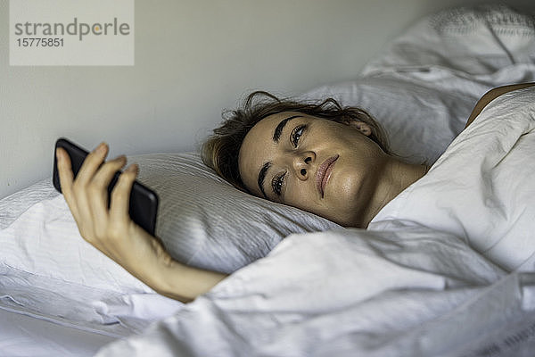 Junge Frau schaut auf dem Bett auf ihr Smartphone