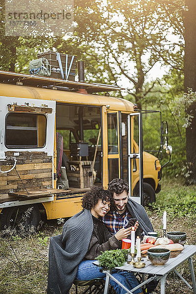 Junges multiethnisches Paar  das sich ein Mobiltelefon teilt  während es beim Camping in eine Decke gewickelt ist