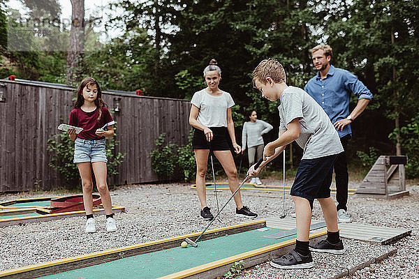 Familie in voller Länge schaut auf einen Jungen  der im Garten Minigolf spielt