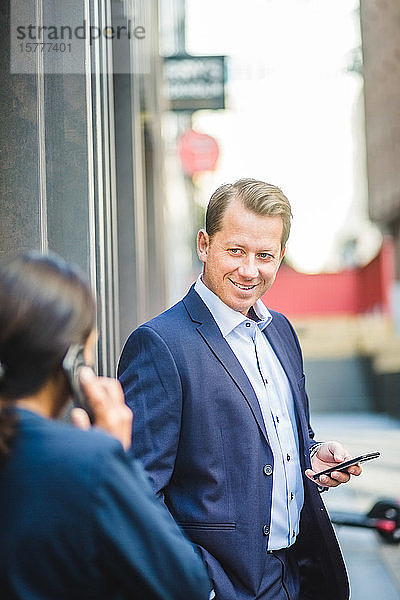 Lächelnder reifer Geschäftsmann mit Smartphone  der einen Kollegen im Freien ansieht