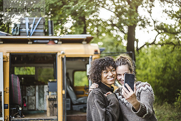 Frau nimmt Selfie mit Freundin gegen Wohnwagen beim Camping im Wald