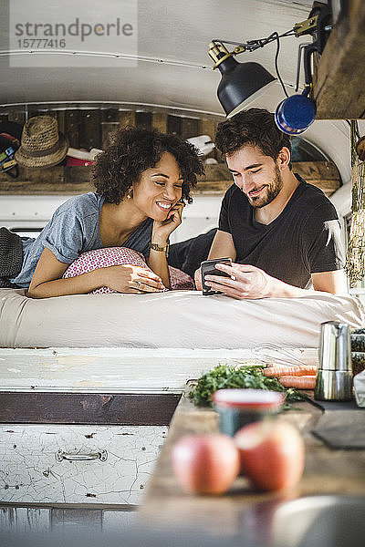 Lächelndes Paar  das sich ein Smartphone teilt  während es im Wohnwagen auf dem Bett liegt