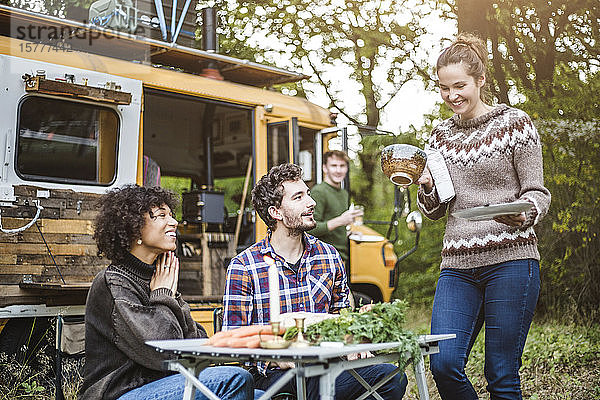 Lächelnde Frau serviert Freunden  die beim Zelten im Wald am Tisch gegen einen Wohnwagen sitzen  Essen