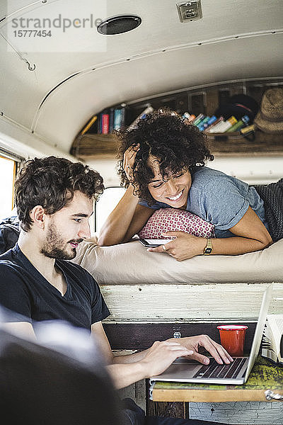 Lächelnde junge Frau im Bett liegend  während sie den Mann mit dem Laptop im Wohnmobil anschaut