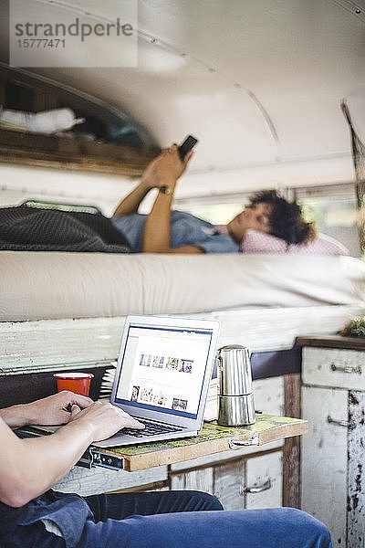 Mittelsektion eines Mannes  der einen Laptop benutzt  durch eine Frau mit Mobiltelefon  die im Wohnmobil auf dem Bett liegt