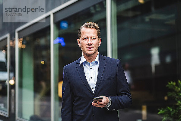 Porträt eines reifen Geschäftsmannes mit Smartphone vor Bürogebäude stehend