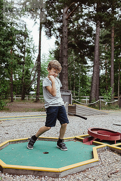 Fröhlicher Junge in voller Länge beim Minigolfspielen im Hinterhof während des Urlaubs
