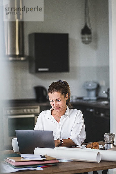 Lächelnde Freiberuflerin benutzt Laptop  während sie zu Hause am Tisch sitzt