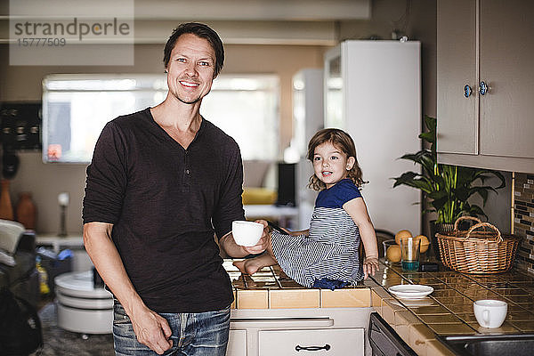 Porträt eines lächelnden reifen Mannes mit Tasse  während die Tochter auf der Küchentheke sitzt