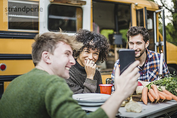 Lächelnder Mann zeigt Freunden  die am Tisch gegen die Karawane sitzen  sein Smartphone