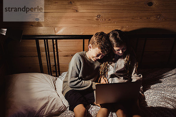 Lächelnde Geschwister mit Laptop  während sie zu Hause im Bett an der Wand sitzen