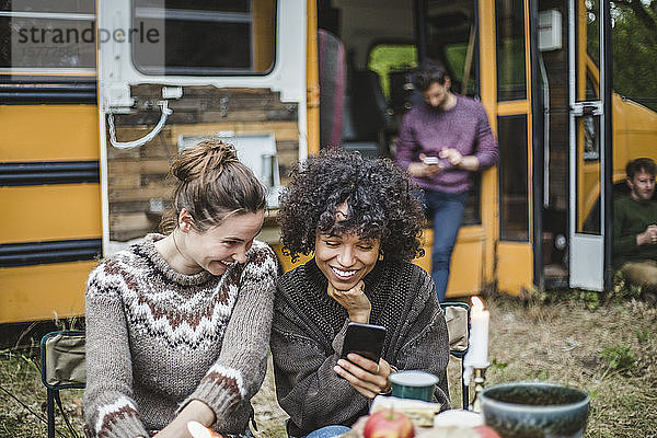 Lächelnde Freundinnen teilen sich ein Smartphone  während sie beim Camping gegen den Wohnwagen sitzen