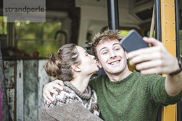 Lächelnder Mann nimmt Selfie vom Handy  während Frau ihn beim Camping küsst