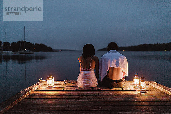 Rückansicht eines Paares  das um eine beleuchtete Laterne am Pier am Meer gegen den Himmel sitzt