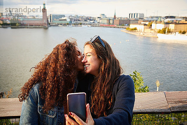 Lächelndes lesbisches Paar  das sich küsst  während es sich mit dem Mobiltelefon gegen das Stadtbild wendet