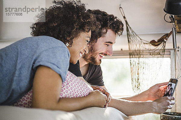 Lächelndes Paar  das im Wohnwagen auf dem Bett liegt und Selfie vom Smartphone nimmt