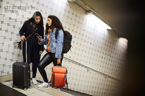 Lesbisches Paar in voller Länge mit Gepäck  das in der U-Bahn steht und ein Mobiltelefon benutzt