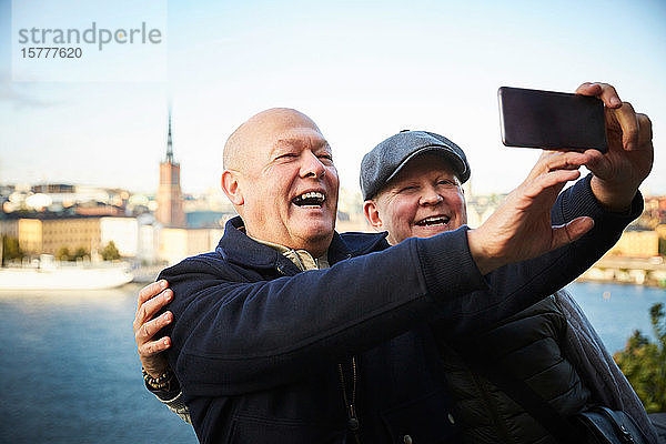 Lächelndes schwules Paar  das sich mit seinem Handy gegen einen Fluss in der Stadt stellt