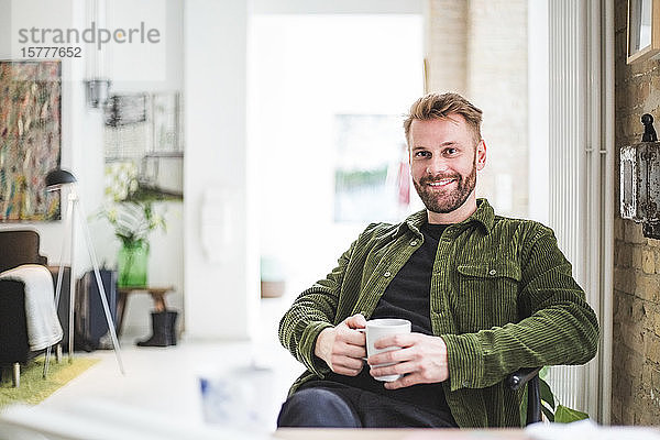 Porträt eines lächelnden männlichen Unternehmers mit Tasse im Heimbüro sitzend