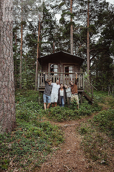 Fröhliche Freunde  die während des Urlaubs mit den Armen um das Haus im Wald stehen