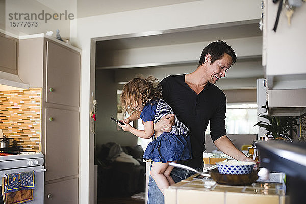 Tochter telefoniert  während der Vater lächelnd in der Küche arbeitet