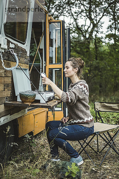 Frau in voller Länge  die während des Zeltens im Wald Kaffee auf dem Herd eines Wohnmobils zubereitet