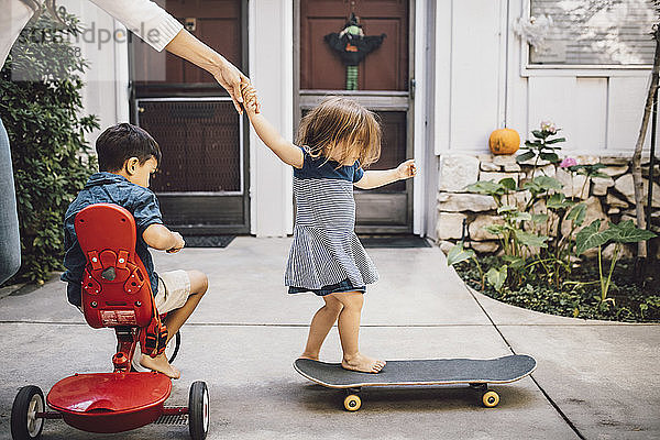 Tochter fährt mit Hilfe der Mutter Skateboard  während der Sohn auf einem Dreirad sitzt