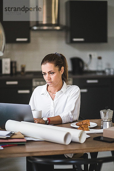 Weibliche Freiberuflerin benutzt Laptop  während sie zu Hause mit Kaffeetasse sitzt