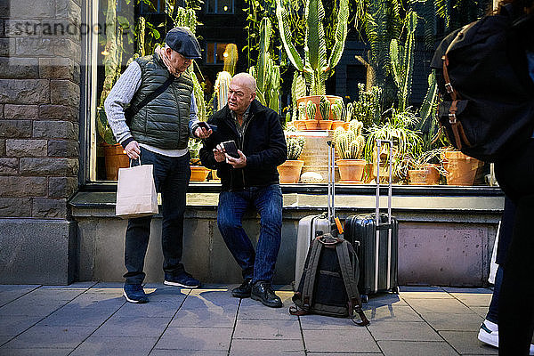 Ein älteres schwules Ehepaar in voller Länge benutzt ein Mobiltelefon  während es nachts vor dem Fabrikladen wartet